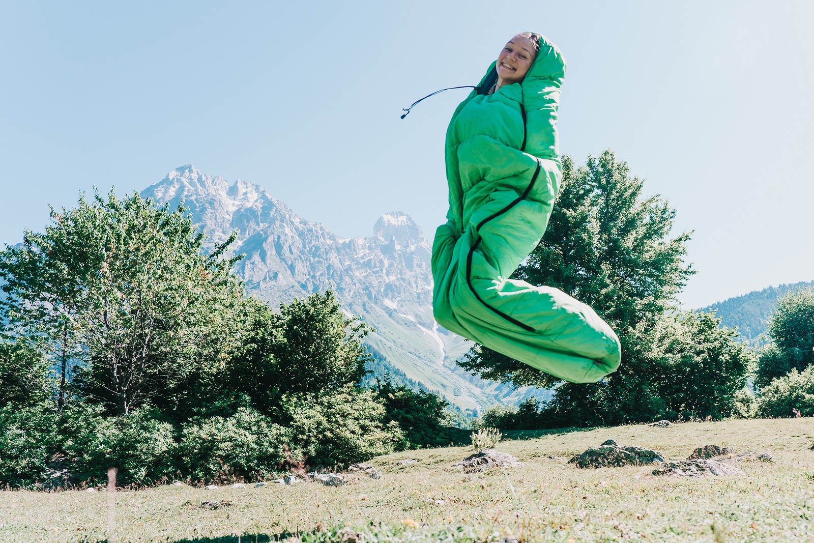 girl in a green sleeping bag and jumping on a background of mountains in sunny weather